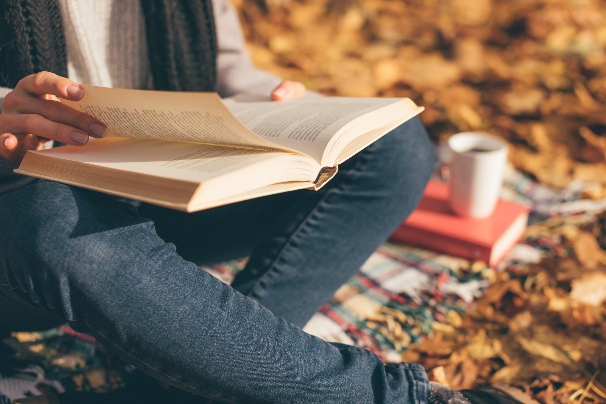 Cropped image of young woman sitting on blanket, reading book and drinking coffee or tea in autumn garden.